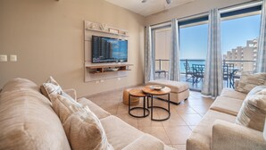 Ein großzügiger Wohnraum mit einem großen Flachbildfernseher an der Wand, einer gemütlichen, beige farbenen Sofagarnitur, einem kleinen Holztisch und einem Balkon mit Blick auf das Meer und die Gebäude.