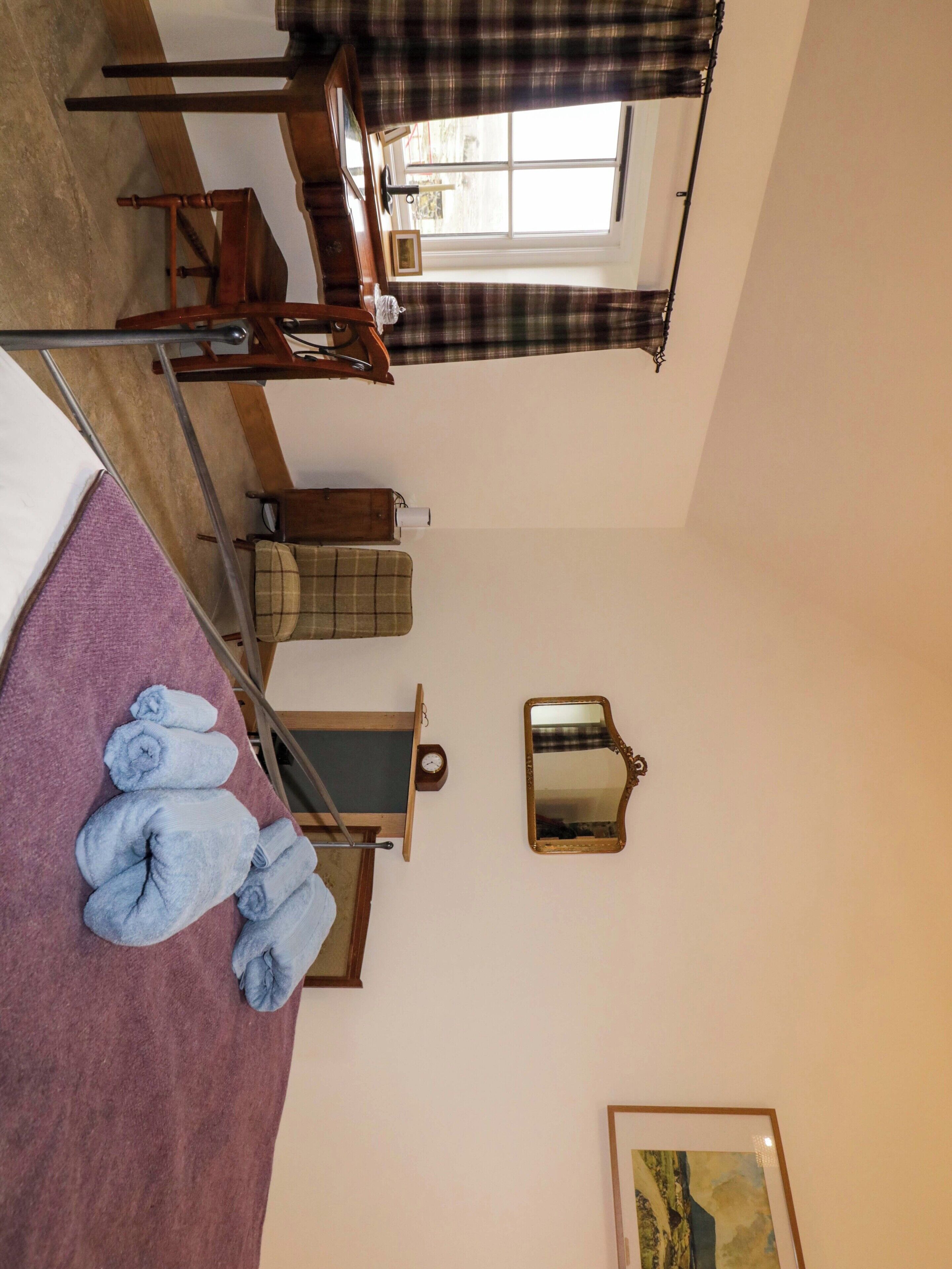 Una habitación con una cama, una silla de madera, una mesita, un espejo y una ventana con cortina escocesa.