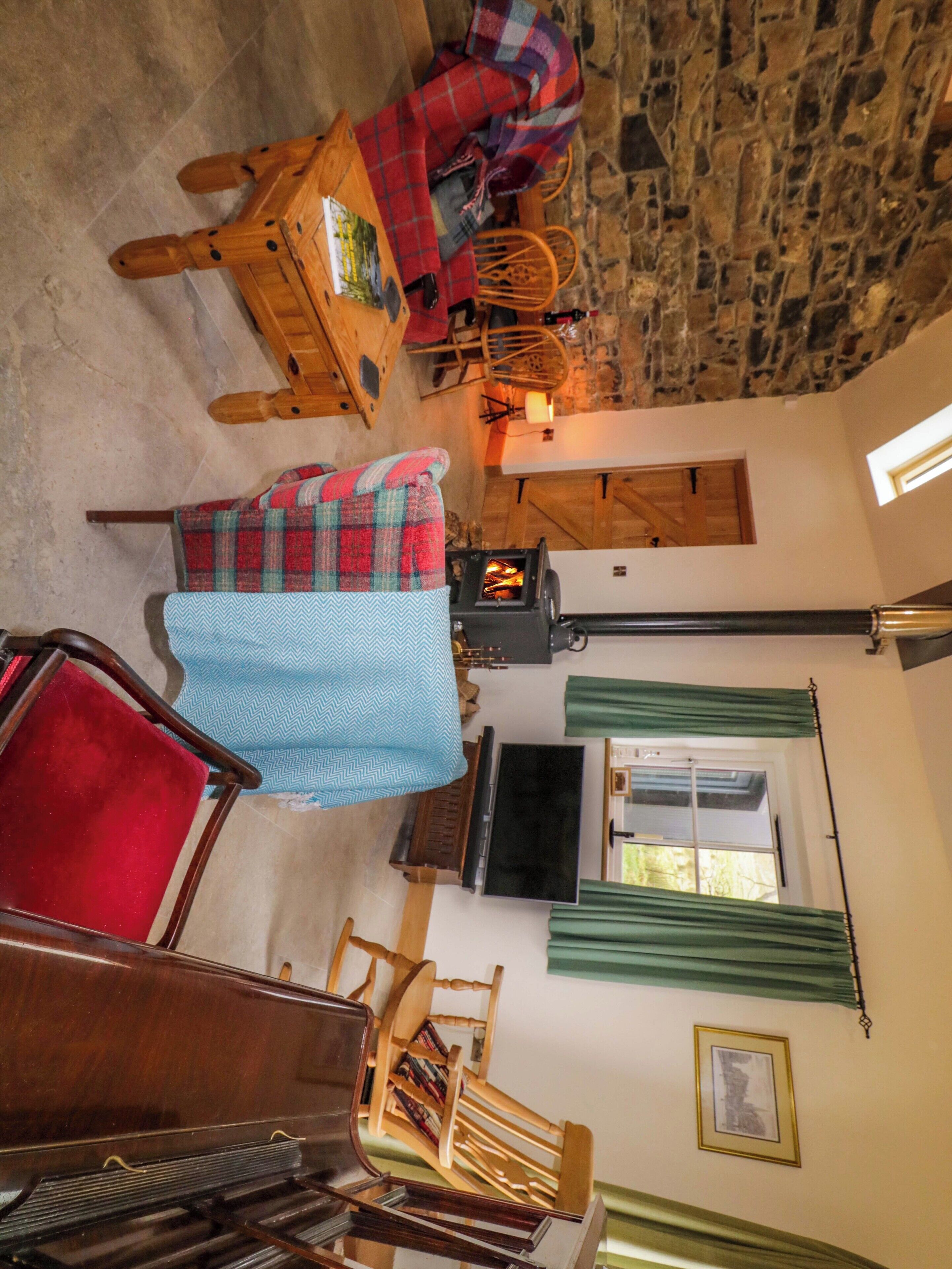 A cozy room with a stone wall, a fireplace, a wooden table with a plaid blanket, a red chair, a wooden bookshelf, and a television.