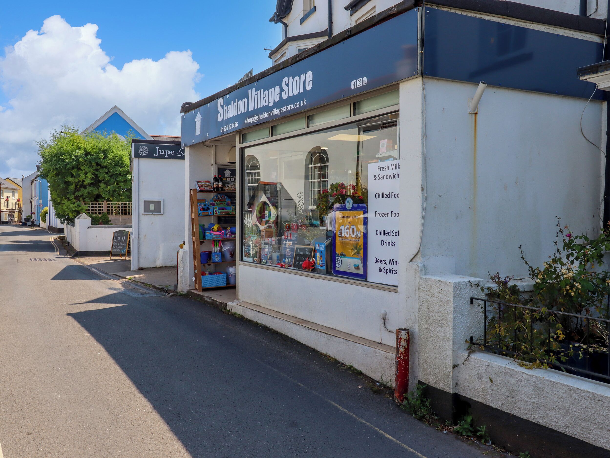 A storefront with a sign reading "Shaldon Village Store" and a display window advertising various products like fresh milk, sandwiches, and frozen food.