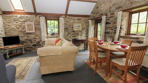 A cozy room with a stone wall, a sofa, a dining table with chairs, and a television on a wooden stand. - The Old Stable (Fishguard)
