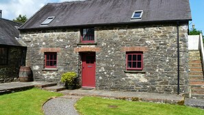 A stone building with a red door, two windows with red frames, and a small garden area with a barrel and a potted plant. - The Old Stable (Fishguard)