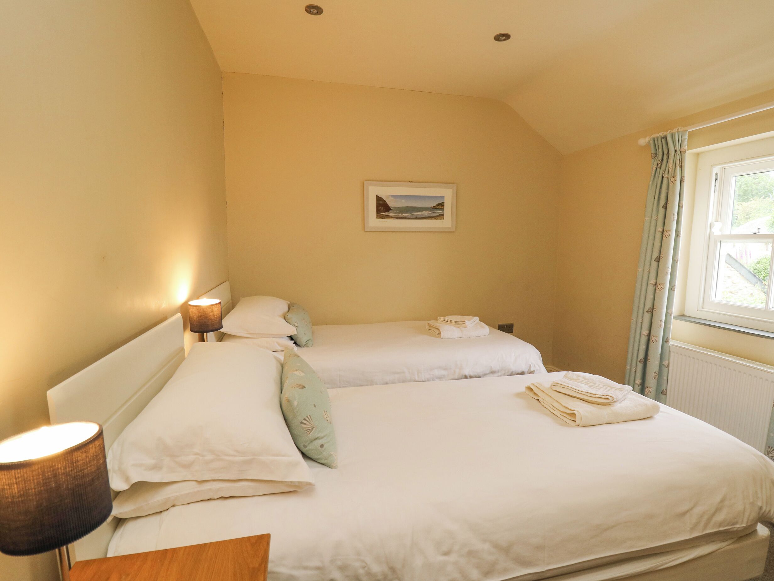 A bedroom with two beds, a bedside lamp, a framed picture on the wall, and a window with curtains.