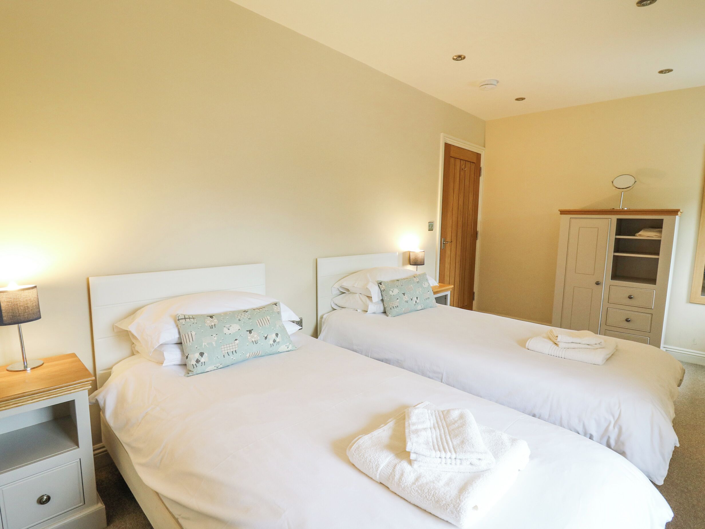 A hotel room with two single beds, a wooden wardrobe, a bedside table with a lamp, and a door leading to another room.