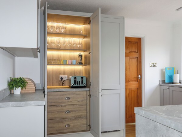 A kitchen with wooden cabinets, a built-in shelf with glasses, a toaster, and a potted plant. - Hafan Deg (Pwllheli)