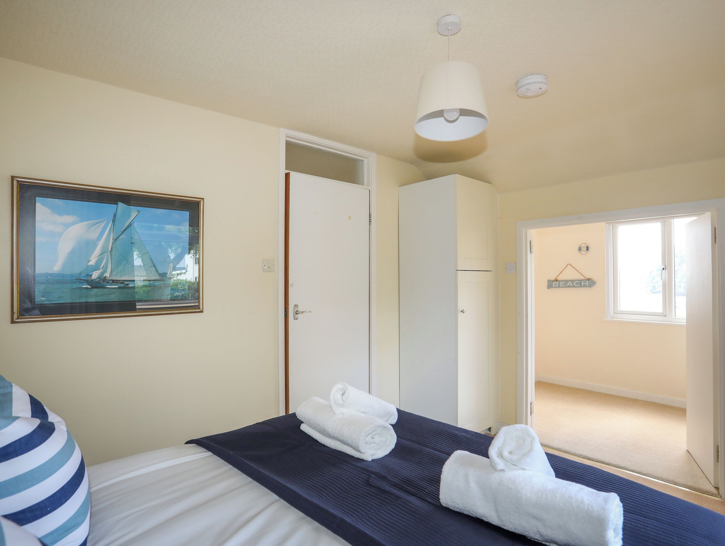 A hotel room with a bed, a framed picture of a sailboat, a wardrobe, a door, and a sign indicating a beach direction.