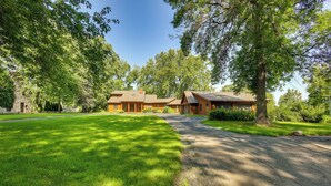 A two-story log cabin with a stone foundation, surrounded by lush greenery and a well-maintained lawn.