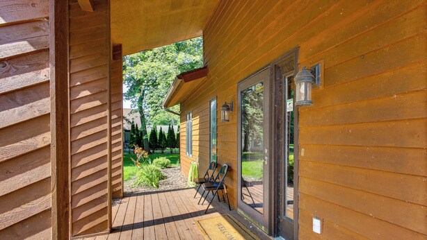 A wooden cabin with a wooden deck, a welcome mat, and two chairs outside.