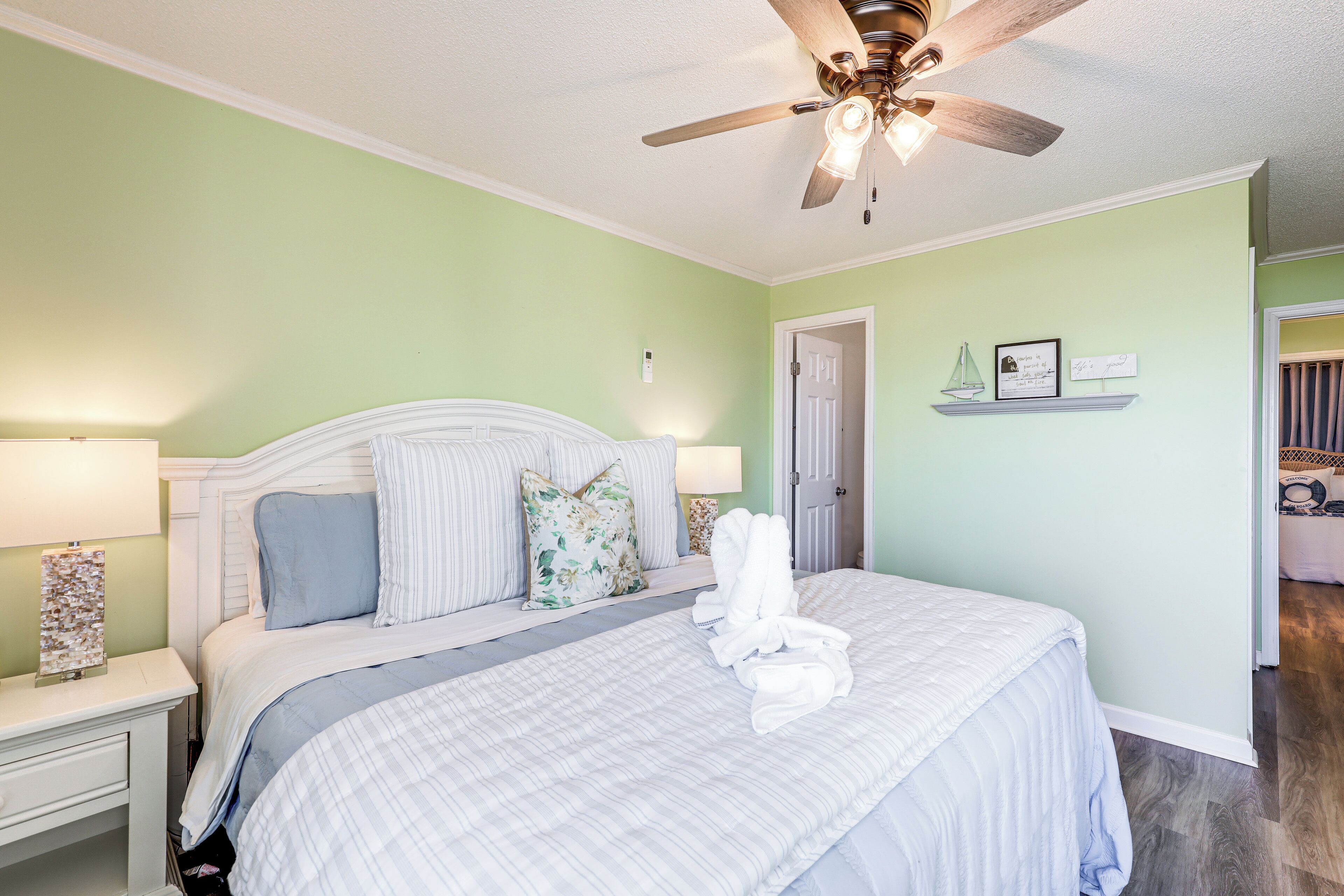 Un lit bien fait, recouvert d’une couvre-lit à rayures blanches et bleues, avec une tête de lit en bois et un ventilateur de plafond au-dessus.