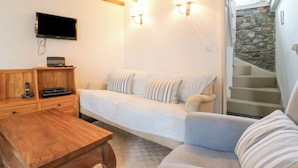 A cozy room with a wooden table, a sofa, a TV mounted on the wall, and a staircase leading to a stone-walled area.
