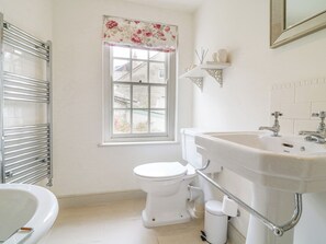 Un baño con inodoro y lavamanos blancos, y una bañera. Hay una ventana con cortina floral y vistas a edificios.