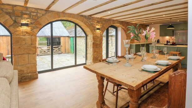 A rustic dining area with a wooden table set for six, stone walls, and a view of the outside.