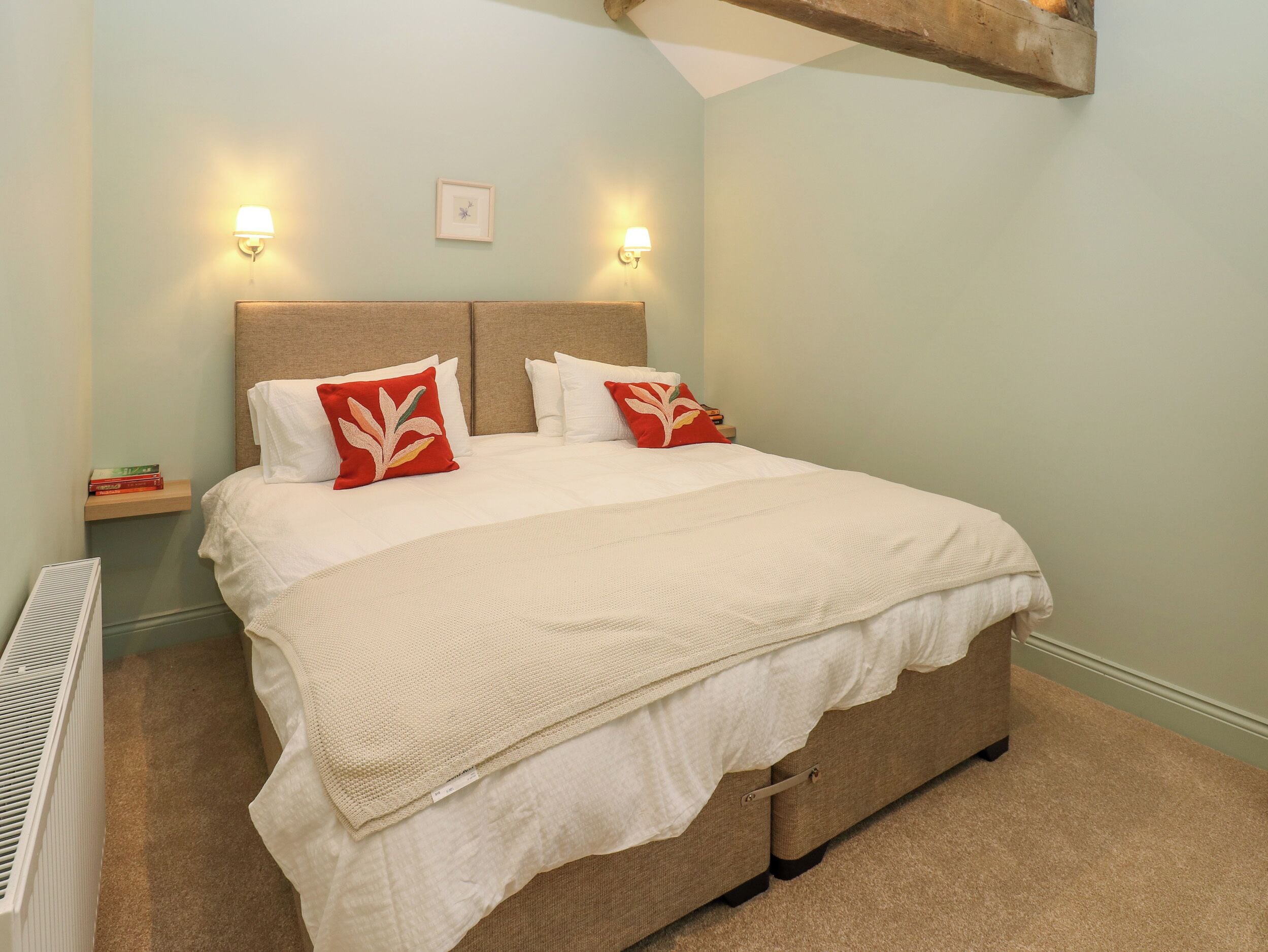 A neatly made bed with white bedding and red decorative pillows, a bedside table with a lamp, and a framed picture on the wall.