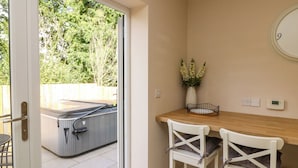 A room with a sliding glass door leading to a patio with a hot tub. There are two white chairs with cushions and a small table with a vase of flowers. - Spicery (Exeter)