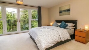 A bedroom with a bed, a nightstand, a lamp, a painting, and a view of greenery through the window. - Spicery (Exeter)