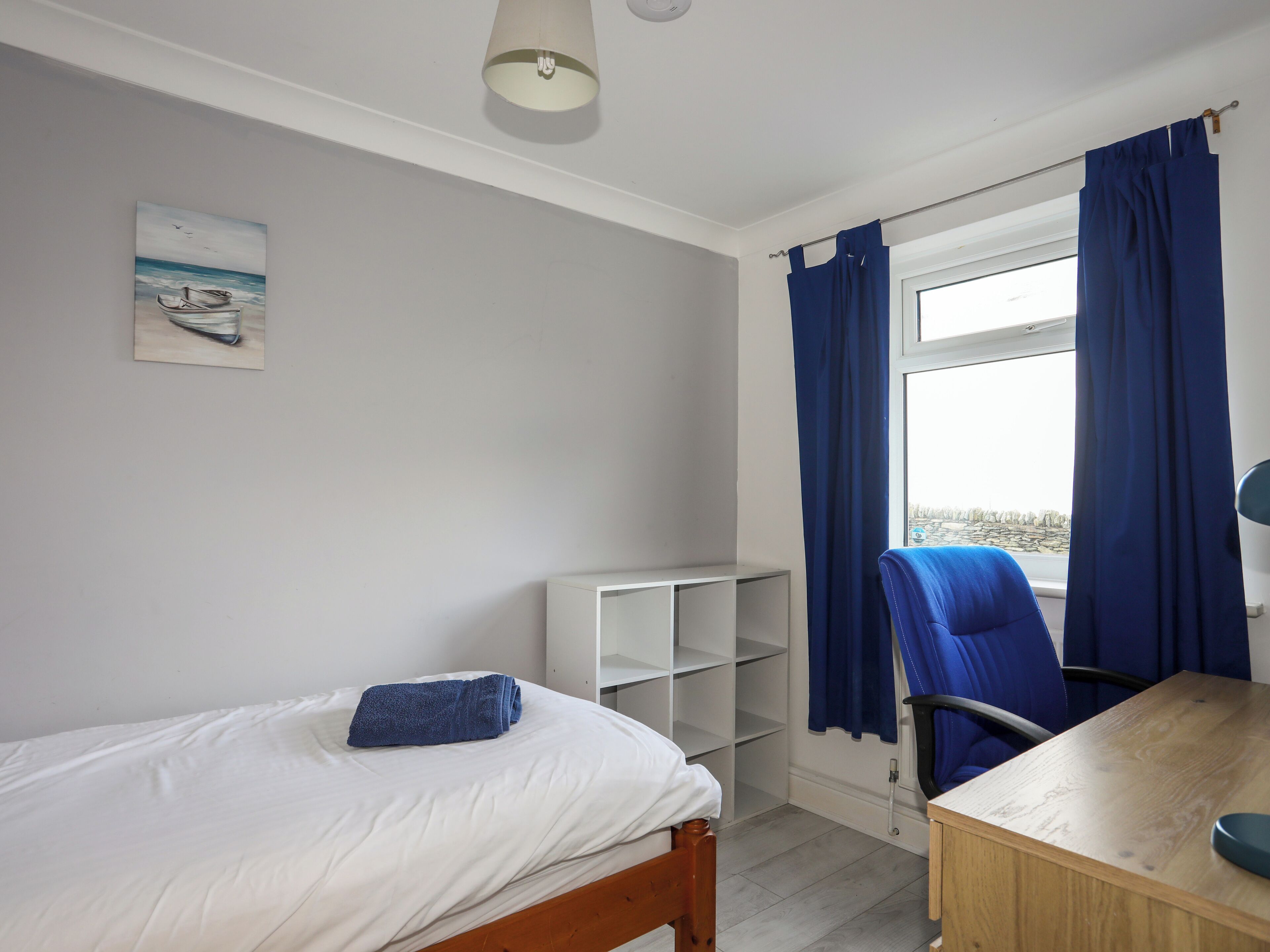 Um quarto com cama, escrivaninha, cadeira, janela com cortinas, estante e um quadro na parede.