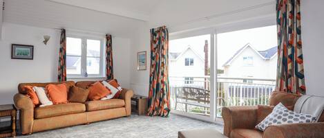 Una sala de estar con un sofá, un reposapiés y una pequeña mesa. Hay ventanas con cortinas, un radiador y un cuadro en la pared.