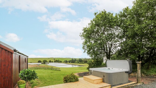 A hot tub with a wooden deck, a wooden shed, and a grassy field with a pond in the background. - Mallard Lodge (Galston)