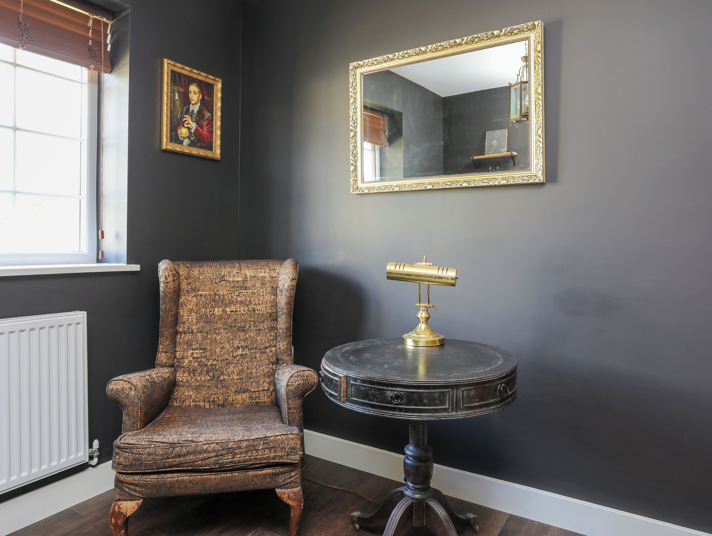 A room with a dark wall, a patterned armchair, a small round table with a brass lamp, and a framed picture.