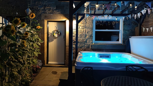 Un jacuzzi bajo una pérgola con luces colgantes, un muro de piedra y una puerta adornada con un adorno navideño.