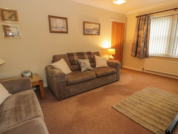 A cozy living room with a brown sofa, a small side table, a lamp, and a window with curtains. - The Barn at Daldorch (Mauchline)