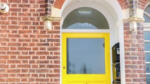 Une porte jaune avec un panneau de verre, encastrée dans un mur de briques surmonté d’une arche blanche.