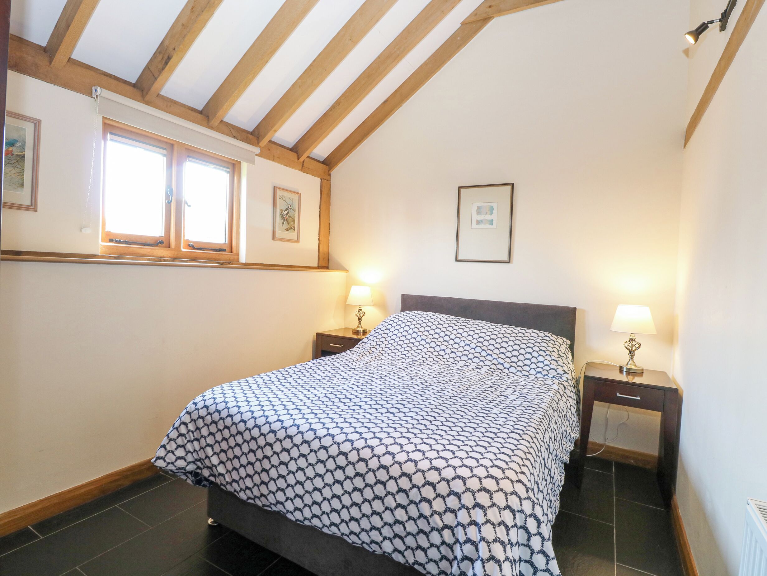 A bedroom with a bed, two bedside tables, a window, and a ceiling with wooden beams.