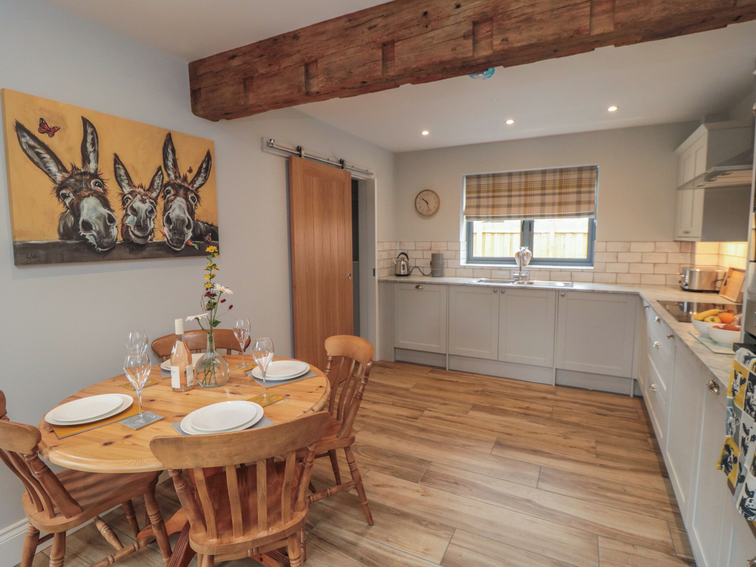 A dining area with a wooden table and chairs, a painting of horses, a kitchen with white cabinets, and a window with a plaid curtain.