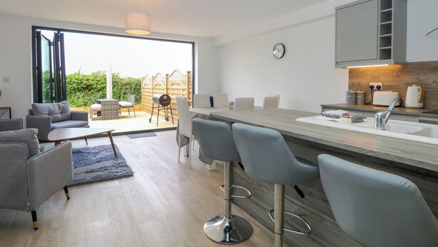 A modern kitchen with a large island, bar stools, and a dining area with a sofa and chairs. - Clavering 1 Bernaleen Cottages (King's Lynn)