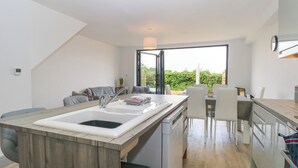 A modern kitchen with a central island, a dining area with chairs, and a view of greenery outside. - Clavering 1 Bernaleen Cottages (King's Lynn)