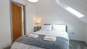 A bedroom with a bed, a nightstand, a lamp, a wooden door, and a skylight. - Clavering 1 Bernaleen Cottages (King's Lynn)