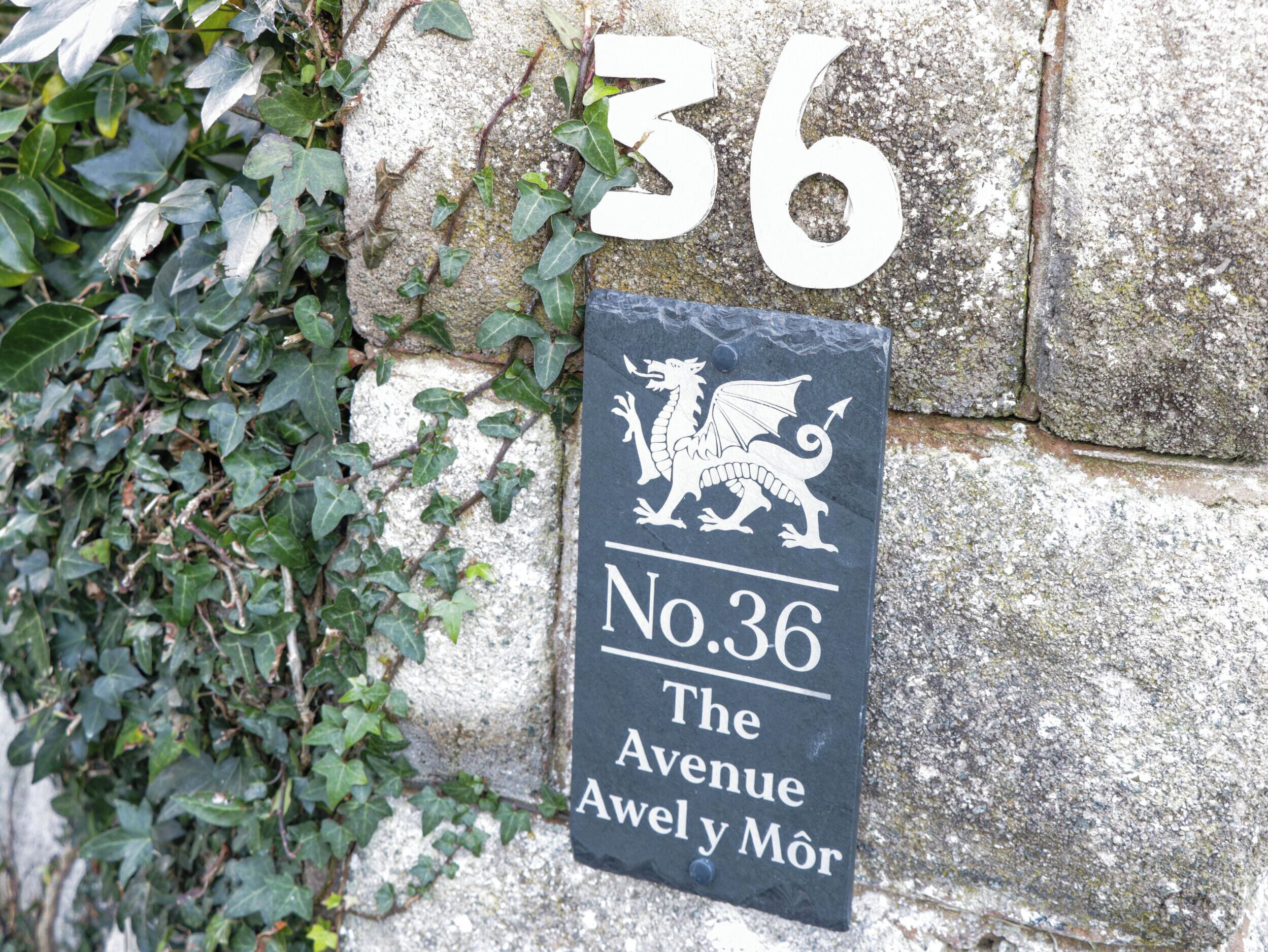 A black sign with white text and a white dragon emblem, indicating 'No.36 The Avenue Avel y Môr'.
