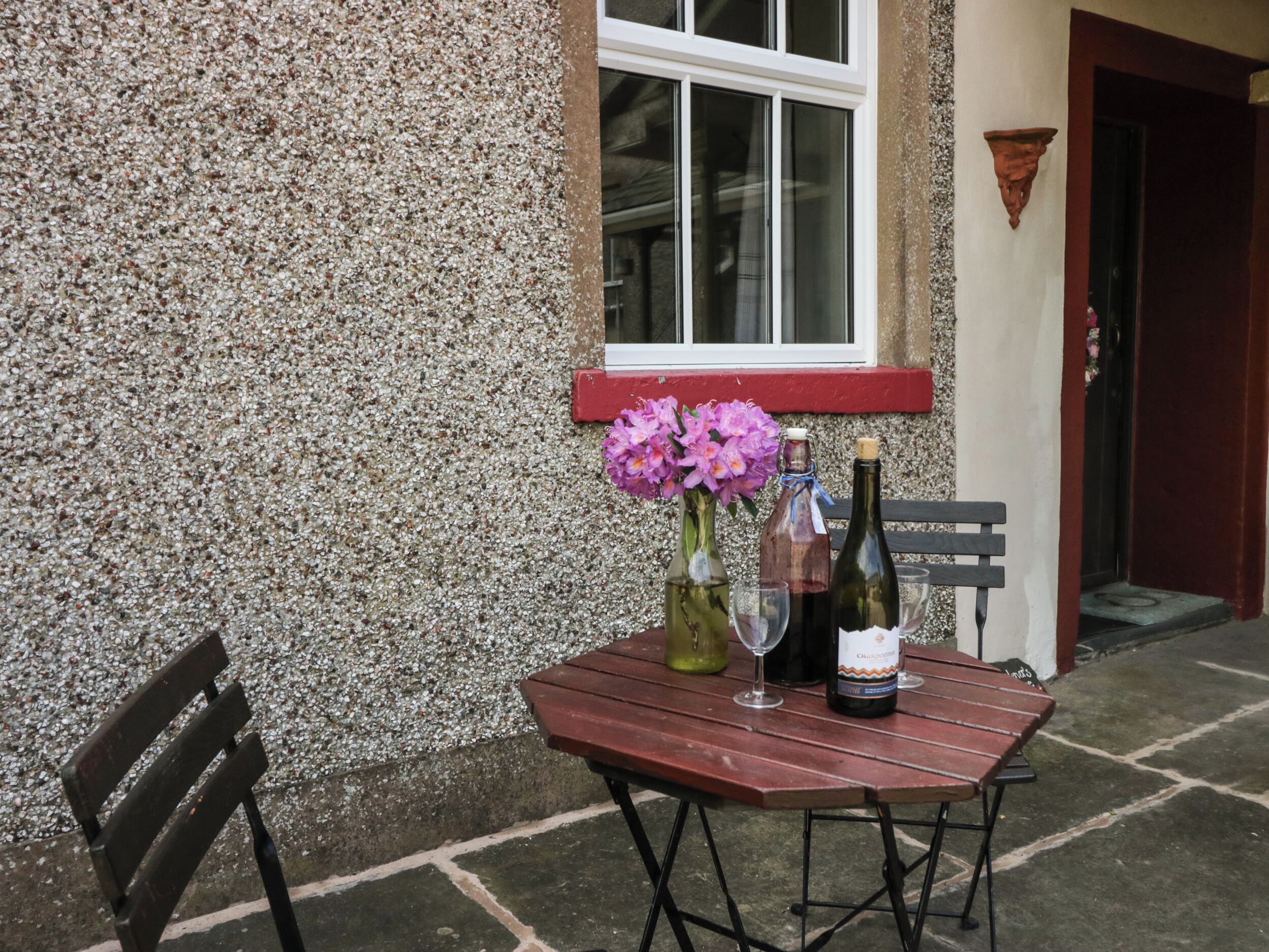 Una mesa de madera con una botella de vino, una copa y un florero con flores en un patio empedrado.
