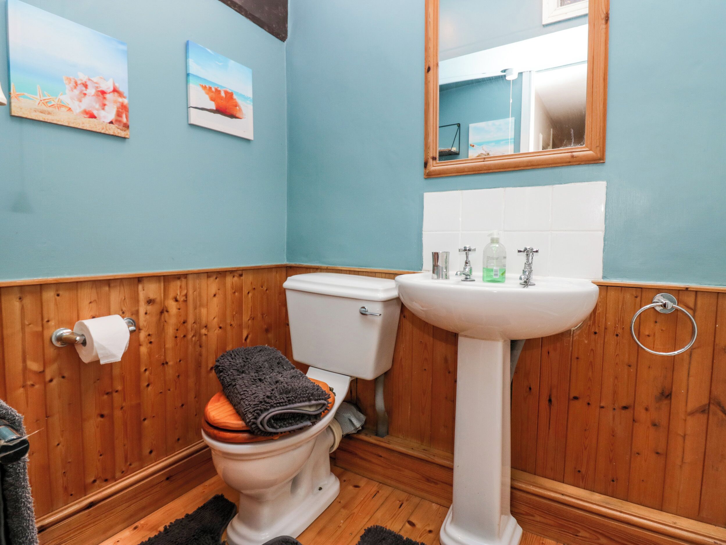 Un baño con suelo de madera, inodoro y lavamanos blancos, y un espejo sobre el lavamanos.