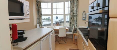 A compact kitchen with a microwave, stove, and sink. There is a dining area with a table and chairs. The room has a bay window with curtains.