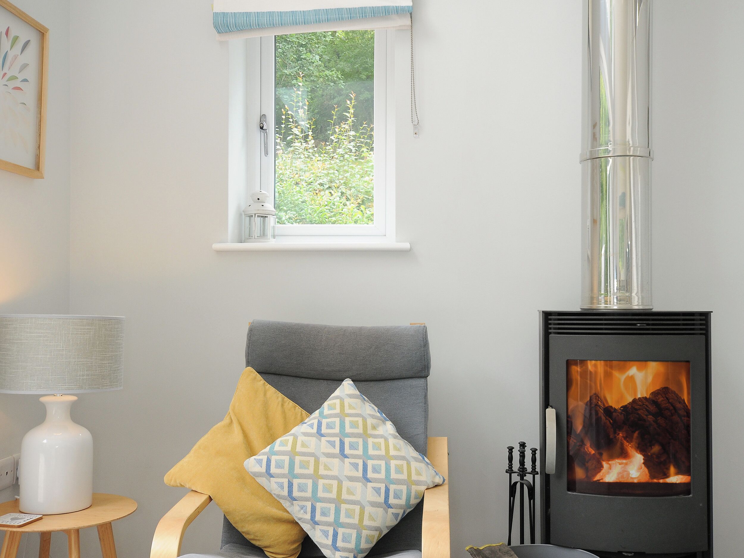 A cozy room with a grey sofa, a patterned throw pillow, a small wooden table with a lamp, and a wood-burning stove.