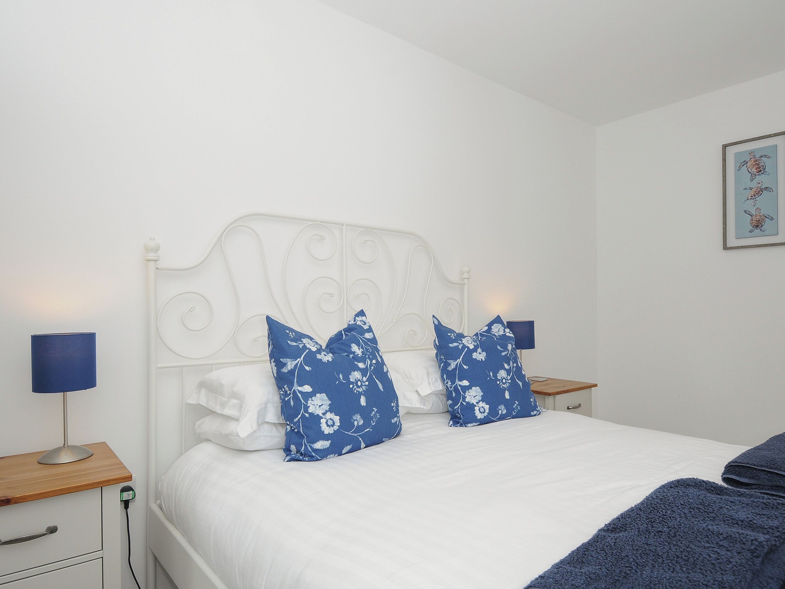A bedroom with a white headboard, two blue floral pillows, a white bedspread, a blue blanket, and a framed picture of a turtle on the wall.