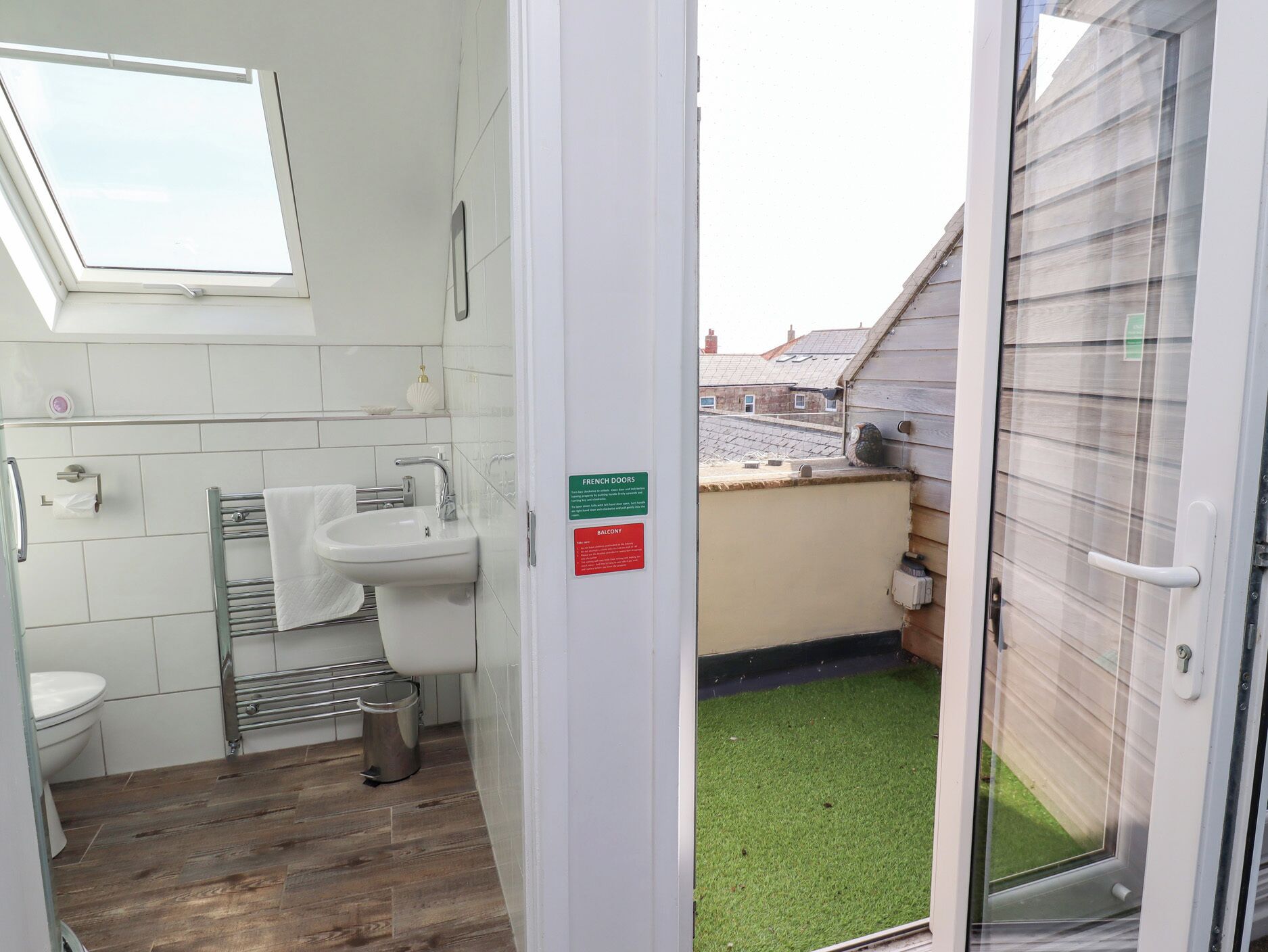 A bathroom with a white sink, a towel rack, and a toilet. There is a skylight above the sink and a door leading to a balcony with artificial grass.