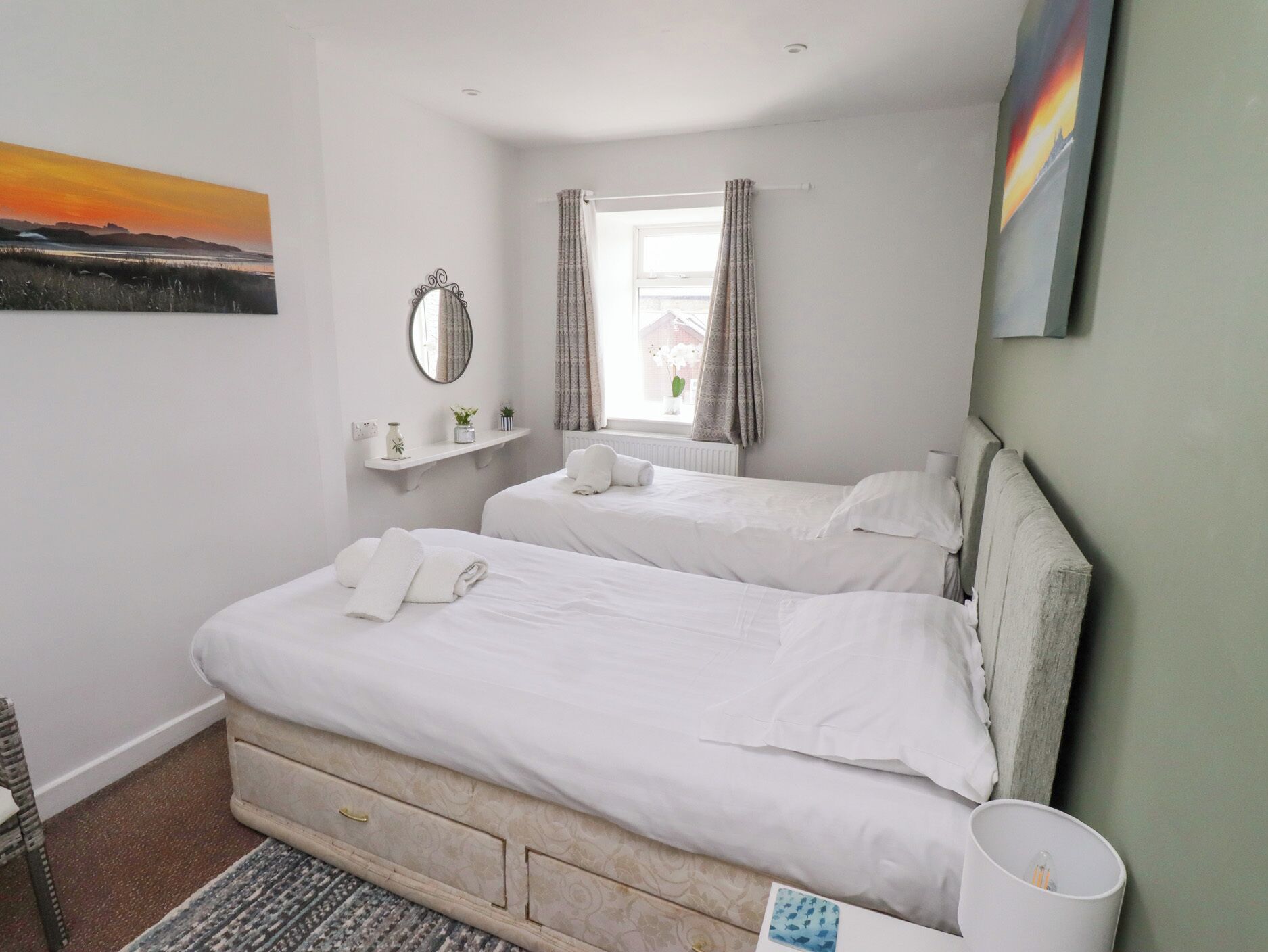 A bedroom with two beds, a window with curtains, a bedside table, a mirror, and a painting on the wall.