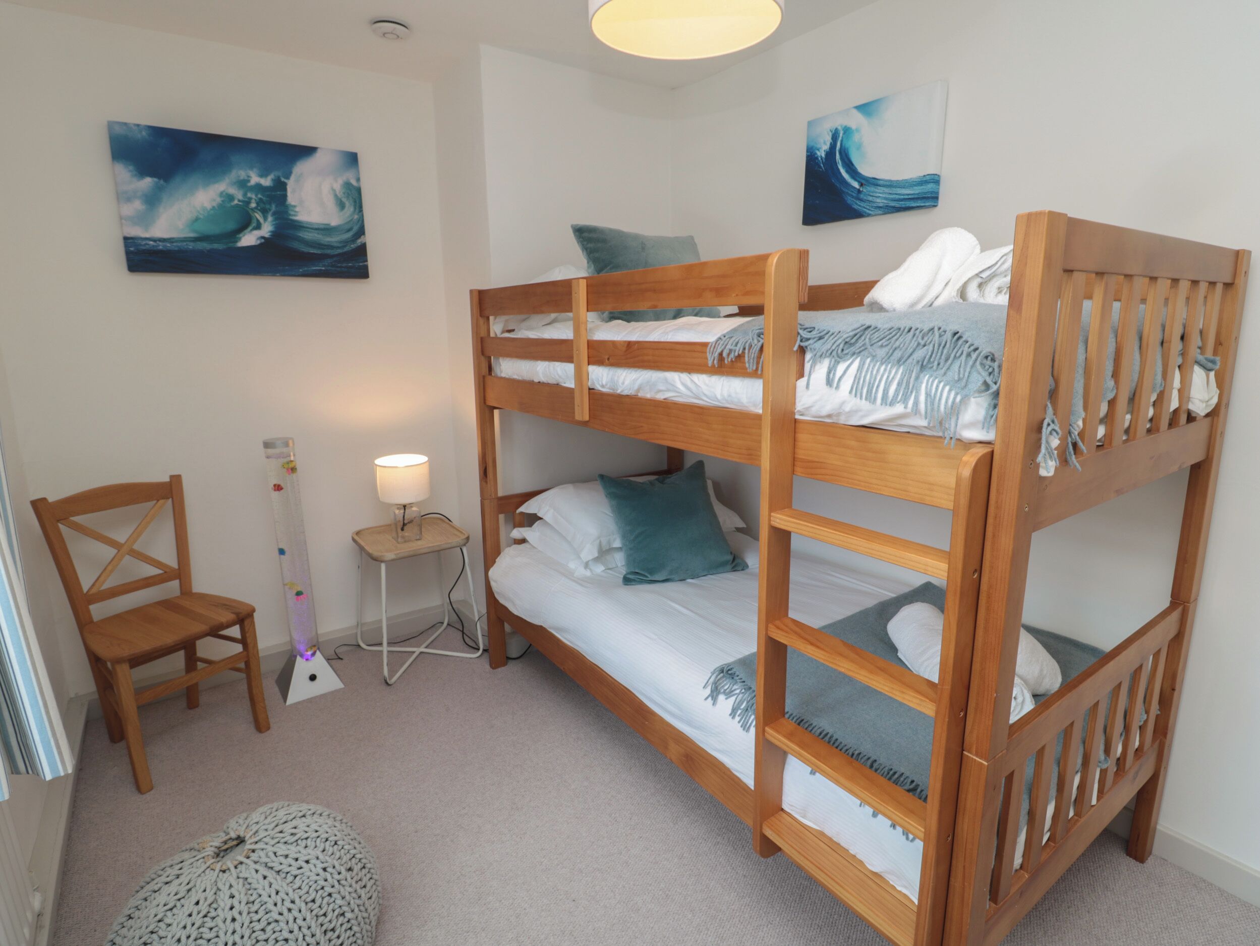 A bunk bed room with a chair, a small table with a lamp, and two wave-themed paintings on the walls.