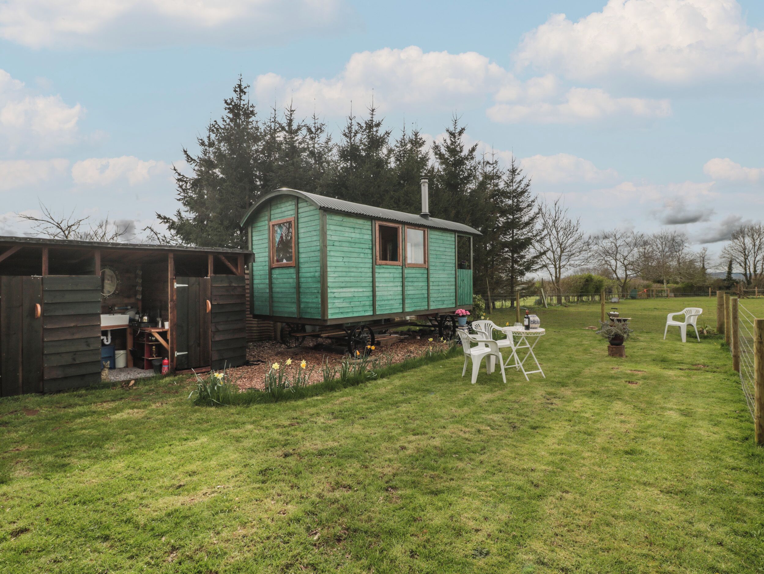 A green wooden house with a small porch, a wooden shed, and a grassy yard with white plastic chairs.