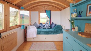 A cozy cabin interior with a wooden ceiling, a bed with patterned pillows, a window with a view of the outdoors, and a shelf with decorative items. - Harri's Hut (Taunton)