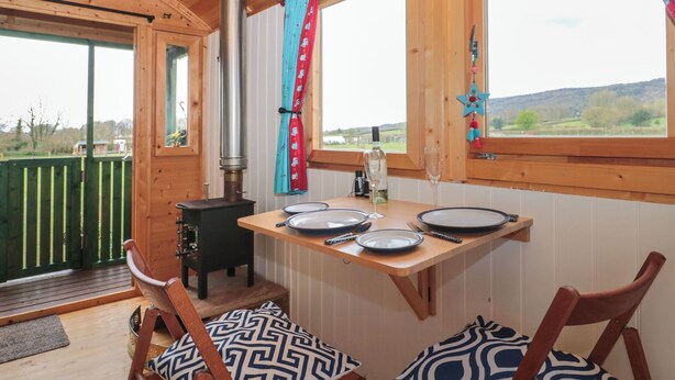 A cozy cabin interior with a dining table set for two, a wood-burning stove, and a view of the outdoors. - Harri's Hut (Taunton)
