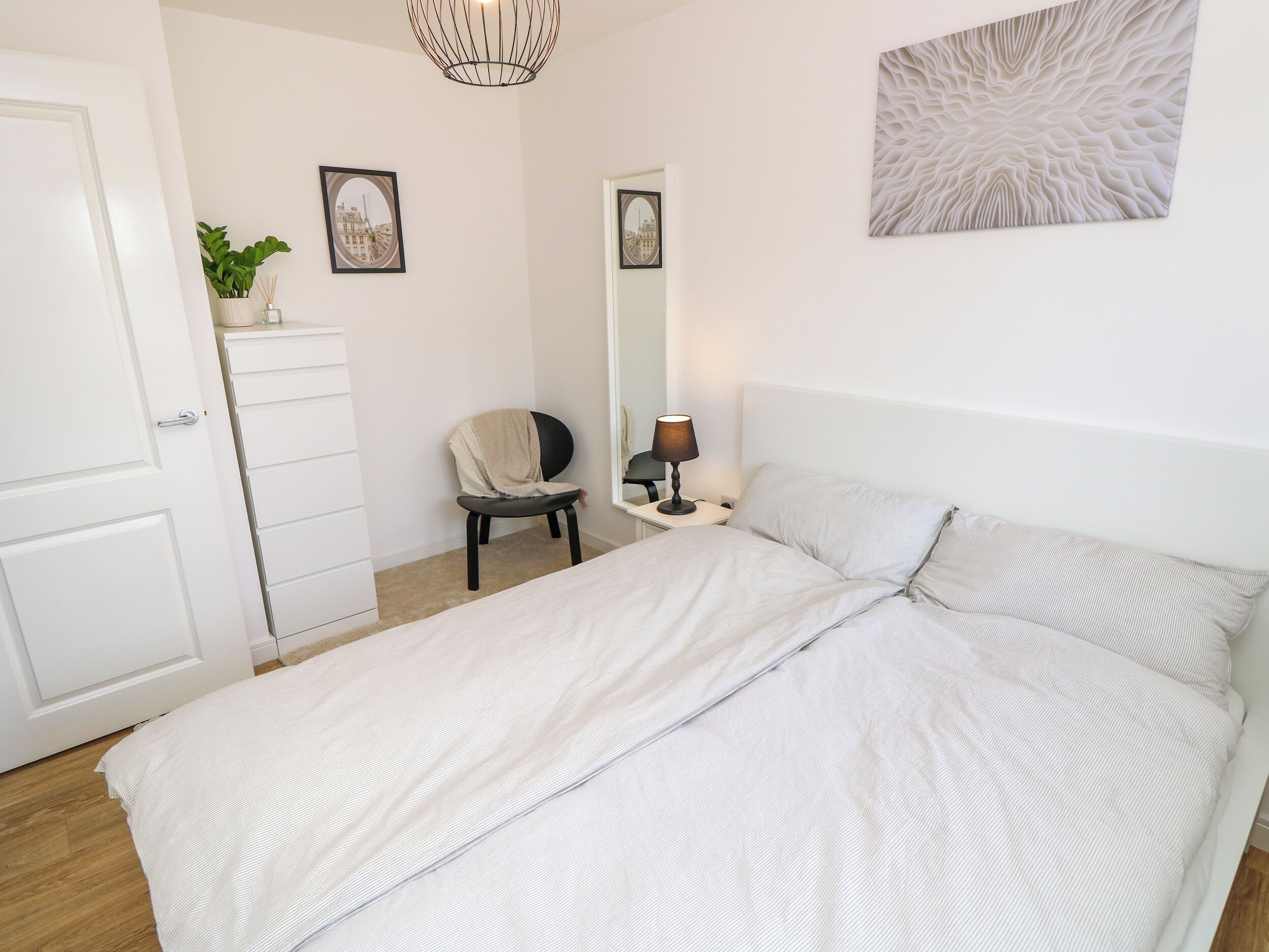 A bedroom with a bed, a chair, a desk with a lamp, a mirror, and a wall-mounted artwork.