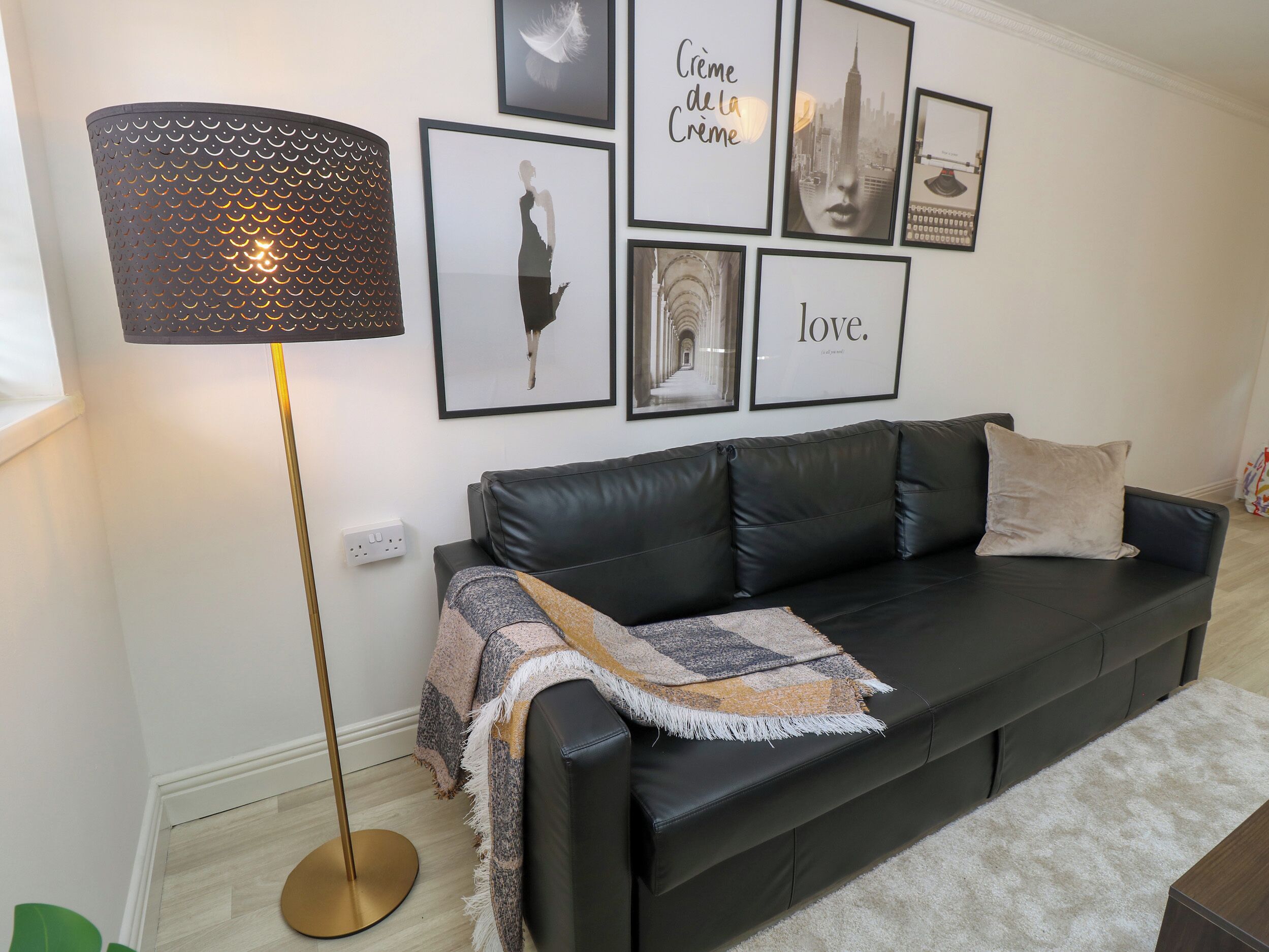 A modern living room with a black leather sofa, a patterned floor lamp, and a series of framed artworks on the wall.
