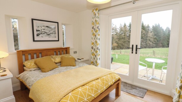 A bedroom with a wooden bed, a bedside table, a framed picture on the wall, and a view of a garden through a sliding door. - Woodland View (Bishop Auckland)