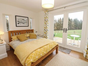 A bedroom with a wooden bed, a bedside table, a framed picture on the wall, and a view of a garden through a sliding door. - Woodland View (Bishop Auckland)