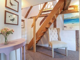 Una escalera con vigas de madera a la vista, un sillón tapizado, obras de arte enmarcadas y un jarrón con flores.