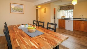 Un comedor con una mesa de madera, sillas y un cuenco de fruta. Se observa una cocina al fondo con armarios de madera y un lavamanos.