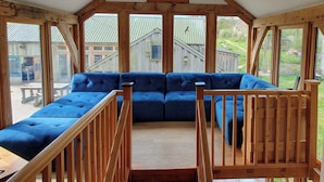 Una habitación con sofá seccional azul, barandales de madera y ventanas amplias que ofrecen vistas a un edificio rústico y vegetación.